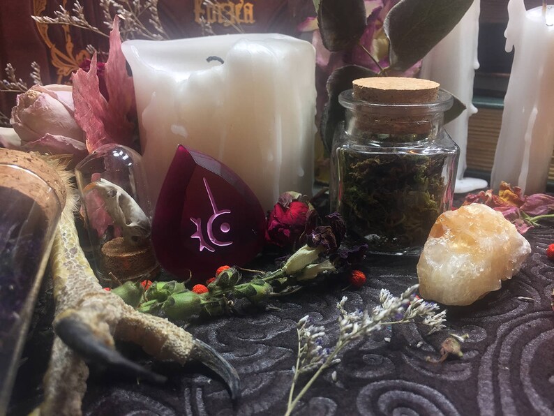 Op de afbeelding: Een close-up van een donkerpaarse, traanvormige kristal met een zilveren symbool erop. De kristal is omringd door gedroogde bloemen, een klein glazen potje met gedroogde kruiden, een witte kaars en een kleine, witte, gladde steen.
