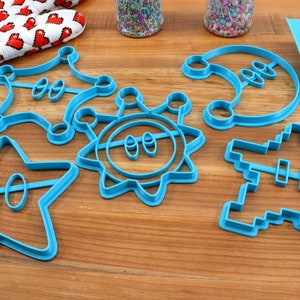 May include: A set of six blue plastic cookie cutters shaped like a star, sun, crescent moon, a pixelated spaceship, and two different shaped crowns.
