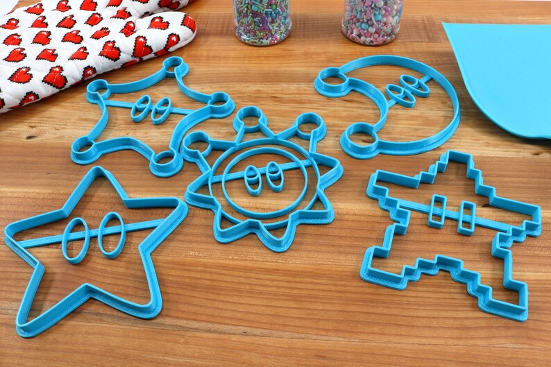 May include: A set of six blue plastic cookie cutters shaped like a star, sun, crescent moon, a pixelated spaceship, and two different shaped crowns.