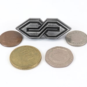 May include: A silver-colored metal brooch with an abstract geometric design, flanked by various coins. The brooch has a repeating pattern and is the focal point of the image. The coins include a penny, a 100 yen coin, and other currency.