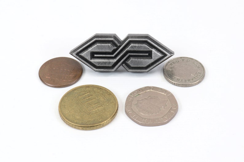 May include: A silver-colored metal brooch with an abstract geometric design, flanked by various coins. The brooch has a repeating pattern and is the focal point of the image. The coins include a penny, a 100 yen coin, and other currency.
