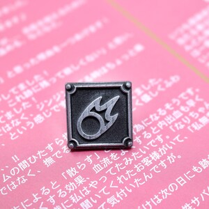 Puede incluir: Un pin de metal plateado con fondo negro y un dise&ntilde;o de llama estilizado. La llama est&aacute; delineada en plata y tiene un c&iacute;rculo negro en el centro.