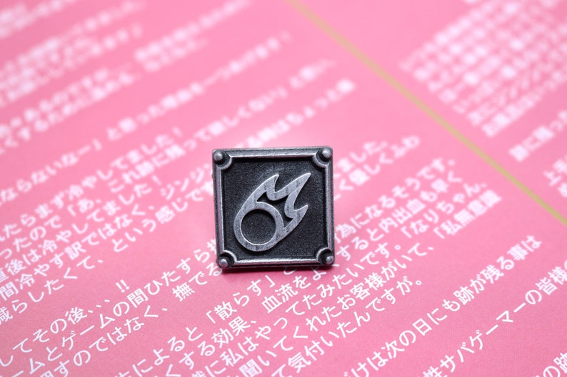 Puede incluir: Un pin de metal plateado con fondo negro y un dise&ntilde;o de llama estilizado. La llama est&aacute; delineada en plata y tiene un c&iacute;rculo negro en el centro.