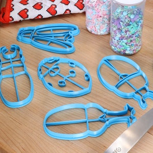 May include: A set of five blue plastic cookie cutters shaped like a ladybug, a beetle, a butterfly, and two other insects. The cutters are on a wooden surface with sprinkles in jars and a blue spatula.