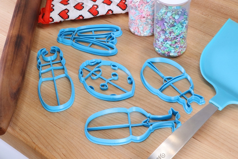 May include: A set of five blue plastic cookie cutters shaped like a ladybug, a beetle, a butterfly, and two other insects. The cutters are on a wooden surface with sprinkles in jars and a blue spatula.