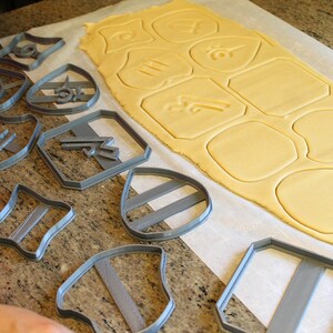 May include: A set of gray plastic cookie cutters in various shapes, including shields, stars, and geometric shapes, arranged on a white surface with a sheet of rolled-out cookie dough.