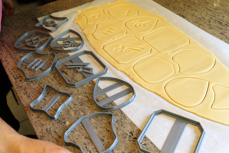 May include: A set of gray plastic cookie cutters in various shapes, including shields, stars, and geometric shapes, arranged on a white surface with a sheet of rolled-out cookie dough.