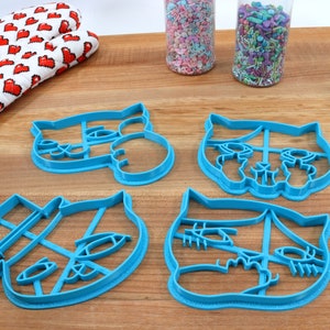 May include: Four blue plastic cat-shaped cookie cutters on a wooden surface.