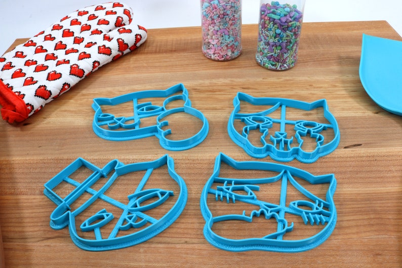 May include: Four blue plastic cat-shaped cookie cutters on a wooden surface.