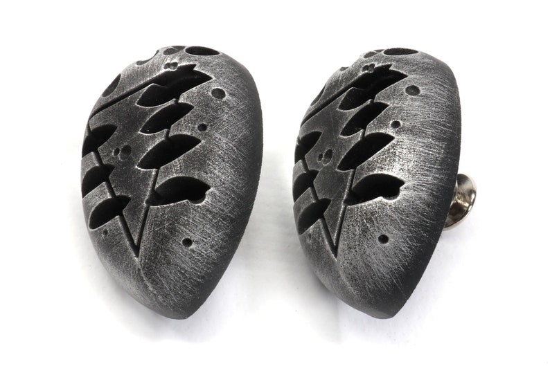 May include: A pair of silver-toned metal earrings shaped like a heart with a cut-out design of a stylized tree branch. The earrings have a textured surface and a pin back.