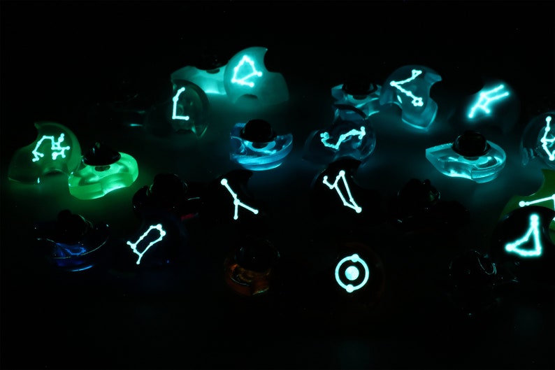 May include: A collection of small, clear glass ornaments with glowing constellations inside. Each ornament features a different constellation, such as Gemini, Cancer, and Leo. The ornaments are arranged on a dark surface, creating a celestial and whimsical display.