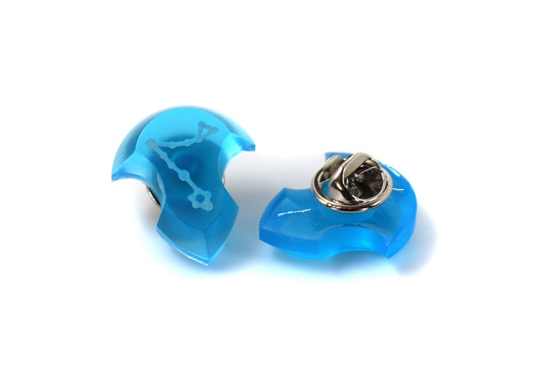 May include: A blue enamel pin with a silver pin back. The pin features a white outline of the constellation Aquarius.