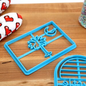 May include: Two blue plastic cookie cutters. One is shaped like the South Carolina state flag with a palmetto tree and crescent moon. The other is shaped like a half circle with a grid pattern.