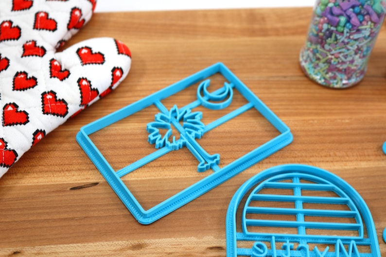 May include: Two blue plastic cookie cutters. One is shaped like the South Carolina state flag with a palmetto tree and crescent moon. The other is shaped like a half circle with a grid pattern.