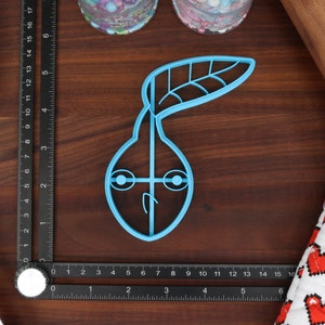 May include: A light blue plastic cookie cutter shaped like a pear with a leaf on top. The pear has two eyes and a mouth.