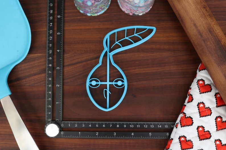May include: A light blue plastic cookie cutter shaped like a pear with a leaf on top. The pear has two eyes and a mouth.