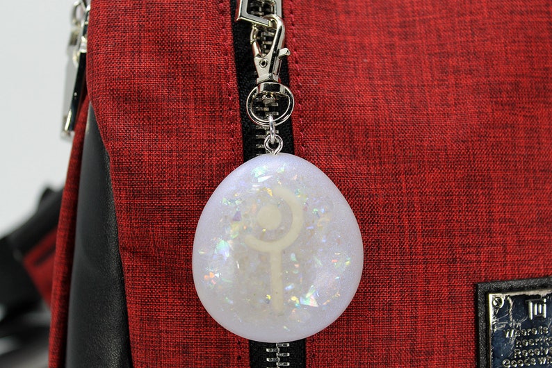 K&ouml;nnte beinhalten: Ein wei&szlig;er eif&ouml;rmiger Anh&auml;nger mit einem silbernen Symbol im Inneren, befestigt an einem silbernen Rei&szlig;verschluss-Zipper. Der Anh&auml;nger befindet sich auf einem roten Stoffhintergrund.