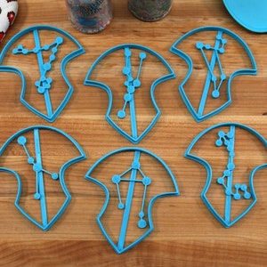 Puede incluir: Seis cortadores de galletas de pl&aacute;stico azul en forma de escudos con dise&ntilde;os de constelaciones en su interior. Los cortadores est&aacute;n sobre una superficie de madera.