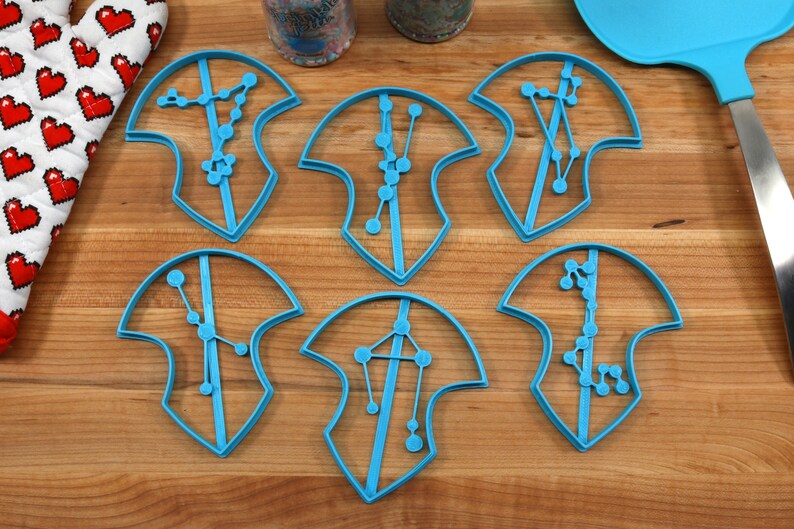 Puede incluir: Seis cortadores de galletas de pl&aacute;stico azul en forma de escudos con dise&ntilde;os de constelaciones en su interior. Los cortadores est&aacute;n sobre una superficie de madera.