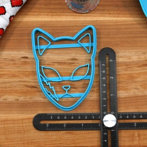 K&ouml;nnte beinhalten: Ein blauer Plastik-Ausstecher in Form eines Katzenkopfes. Der Ausstecher ist etwa 5 cm hoch und hat ein detailliertes Design mit Streifen und einem Blitz.