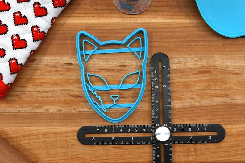 K&ouml;nnte beinhalten: Ein blauer Plastik-Ausstecher in Form eines Katzenkopfes. Der Ausstecher ist etwa 5 cm hoch und hat ein detailliertes Design mit Streifen und einem Blitz.