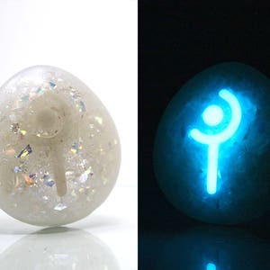 K&ouml;nnte beinhalten: Ein wei&szlig;er Stein mit einem leuchtend blauen Symbol darauf. Das Symbol ist eine stilisierte Strichm&auml;nnchenfigur mit einer geschwungenen Linie &uuml;ber dem Kopf. Der Stein ist eine beliebte Wahl f&uuml;r Fans von Fantasy- und Rollenspielen.