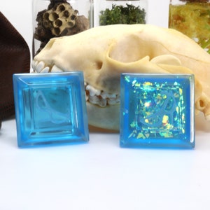 May include: Two square blue resin dice with a white design inside. The dice are on a white surface with a skull and other objects in the background.