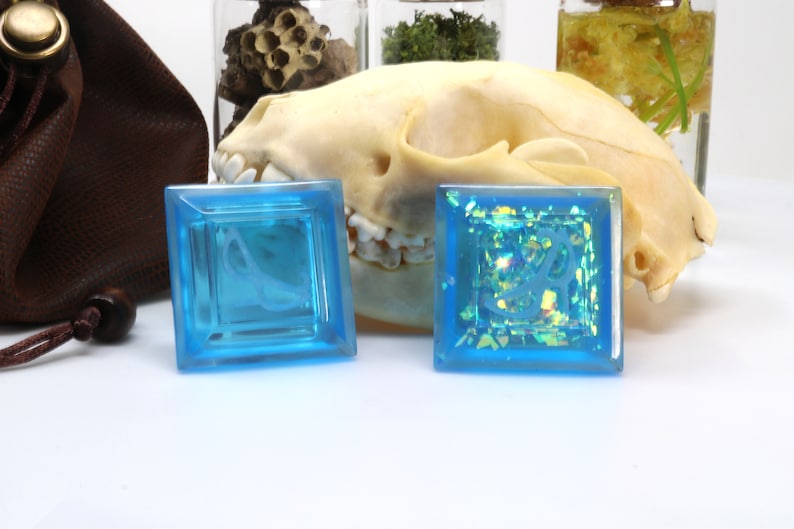 May include: Two square blue resin dice with a white design inside. The dice are on a white surface with a skull and other objects in the background.