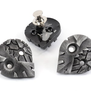May include: Three silver metal buttons with a textured, cracked design. The buttons have two holes each and are shaped like a teardrop. One button has a silver pin attached to the back.