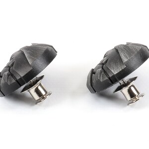 May include: Two black metal buttons with a textured design and a silver pin on the back.