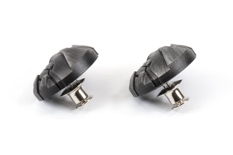 May include: Two black metal buttons with a textured design and a silver pin on the back.