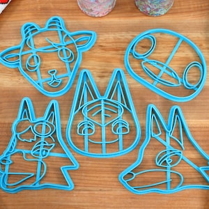May include: Five blue plastic cookie cutters shaped like animal faces. The cutters are on a wooden surface.