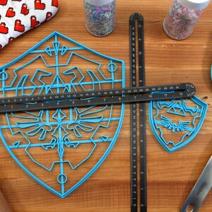 May include: Two blue plastic cookie cutters shaped like the Triforce symbol from the Legend of Zelda video game series. The larger cutter has a grid pattern and the smaller cutter is solid. A black ruler is placed on top of the larger cutter.