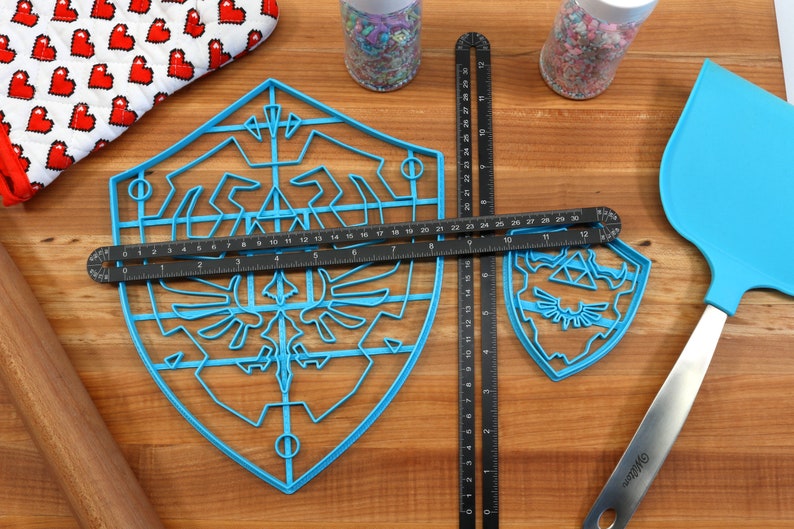 May include: Two blue plastic cookie cutters shaped like the Triforce symbol from the Legend of Zelda video game series. The larger cutter has a grid pattern and the smaller cutter is solid. A black ruler is placed on top of the larger cutter.