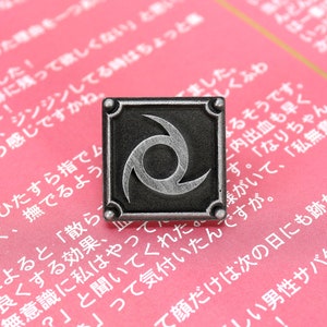 May include: A silver-coloured pin with a black background and a stylised silver swirl design. The pin is on a pink surface with Japanese text.