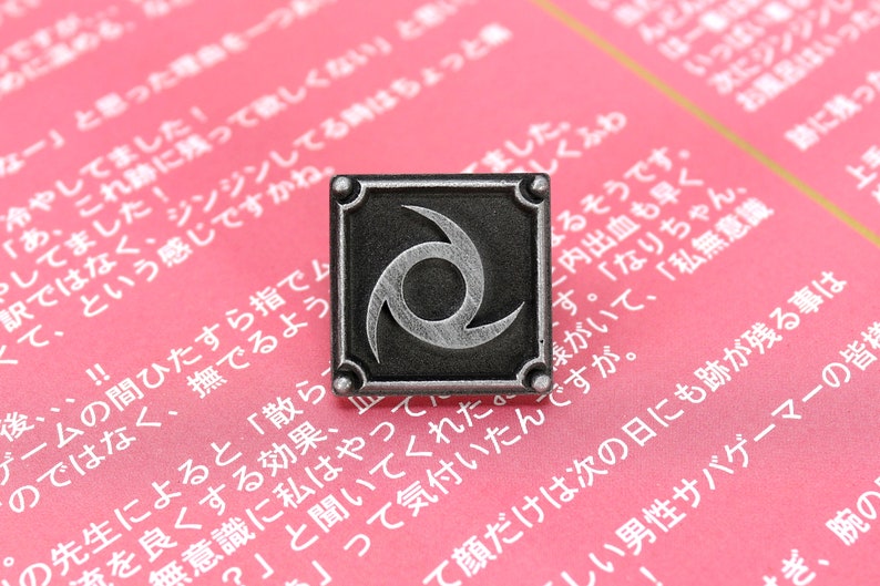May include: A silver-coloured pin with a black background and a stylised silver swirl design. The pin is on a pink surface with Japanese text.