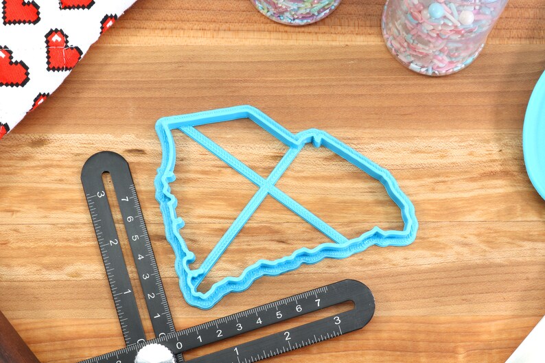 May include: A blue plastic cookie cutter shaped like the state of South Carolina. The cutter is on a wooden surface with a ruler and other baking supplies.