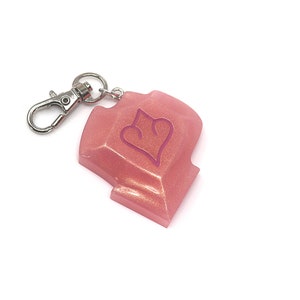 May include: A pink, glittery resin keychain with a silver-colored clasp. The keychain has a shield shape with a heart design in the center. The background is white.