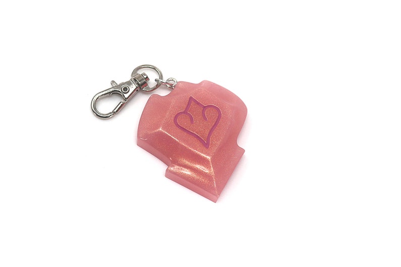 May include: A pink, glittery resin keychain with a silver-colored clasp. The keychain has a shield shape with a heart design in the center. The background is white.