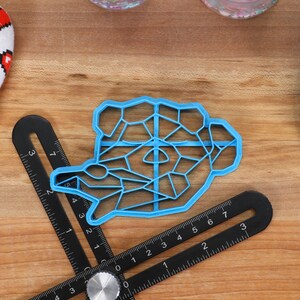 May include: A blue plastic cookie cutter shaped like a geometric animal. The cutter is on a wooden surface with a ruler and a blue silicone mat.