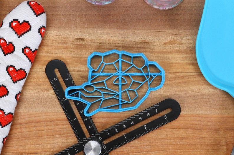May include: A blue plastic cookie cutter shaped like a geometric animal. The cutter is on a wooden surface with a ruler and a blue silicone mat.