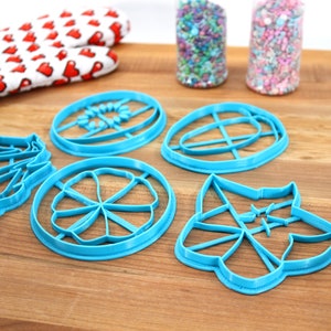 May include: Five blue plastic cookie cutters on a wooden surface. The cutters are shaped like a flower, a circle with a cross inside, a leaf, a star, and a circle with a flower inside.