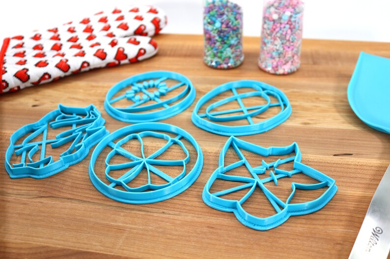 May include: Five blue plastic cookie cutters on a wooden surface. The cutters are shaped like a flower, a circle with a cross inside, a leaf, a star, and a circle with a flower inside.
