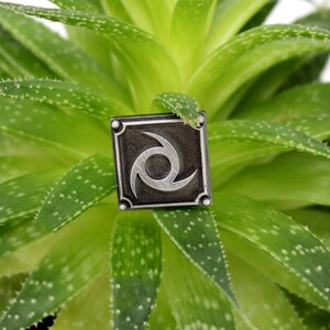 May include: A silver metal pin with a black background and a silver swirl design. The pin is sitting on a green succulent plant.