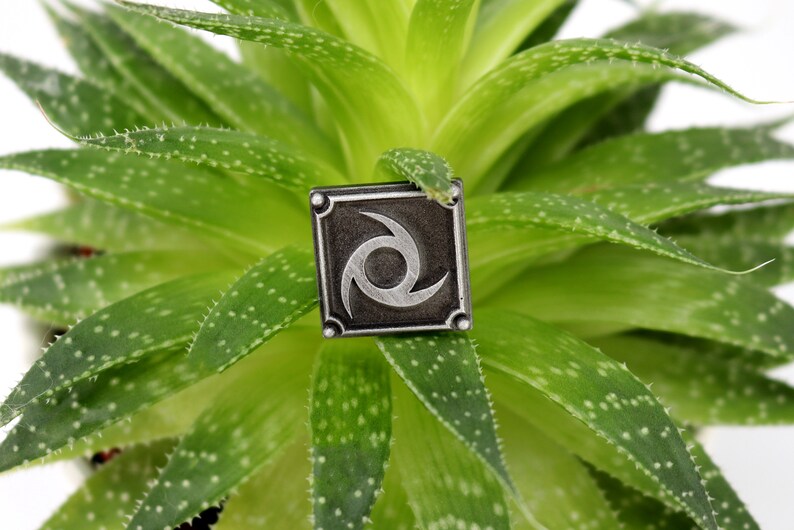 May include: A silver metal pin with a black background and a silver swirl design. The pin is sitting on a green succulent plant.