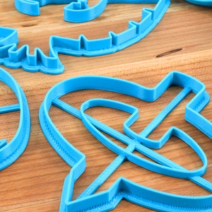May include: Close-up of several bright blue plastic cookie cutters in various shapes, including a dinosaur, a circle, and an abstract design. The cutters are arranged on a light brown wooden surface.