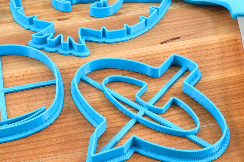 May include: Close-up of several bright blue plastic cookie cutters in various shapes, including a dinosaur, a circle, and an abstract design. The cutters are arranged on a light brown wooden surface.