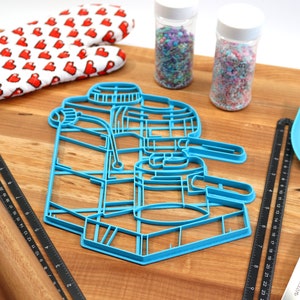 May include: Blue plastic cookie cutter shaped like an espresso machine on a wooden surface with a rolling pin, two jars of sprinkles, a spatula, and a ruler.