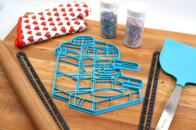 May include: Blue plastic cookie cutter shaped like an espresso machine on a wooden surface with a rolling pin, two jars of sprinkles, a spatula, and a ruler.