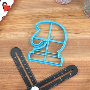 May include: A blue plastic cookie cutter shaped like a chess knight. The cutter is on a wooden surface with a ruler and a container of sprinkles.